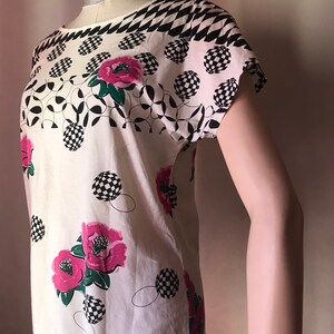 May include: White short-sleeved shirt with a black and white checkered pattern at the neckline and pink flowers with green leaves on a white background.