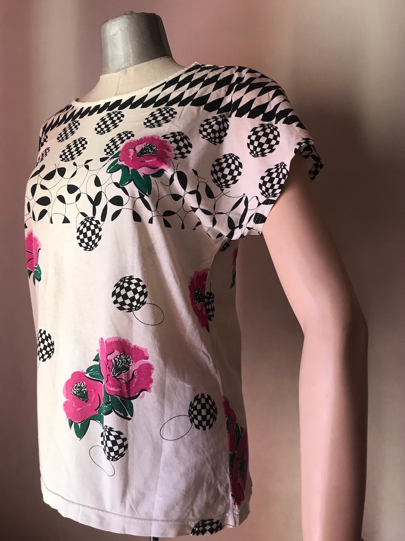May include: White short-sleeved shirt with a black and white checkered pattern at the neckline and pink flowers with green leaves on a white background.