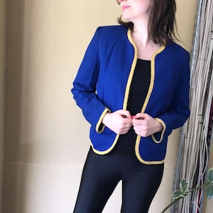 May include: A royal blue blazer with yellow trim. The jacket has a round neckline and long sleeves. The trim is dotted with small yellow circles. The person in the image is wearing black leggings and orange sunglasses.
