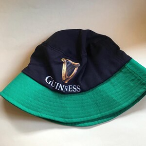 Op de afbeelding: Een zwarte en groene bucket hat met het Guinness-logo en een harpdesign. Het logo luidt "GUINNESS" in witte tekst.