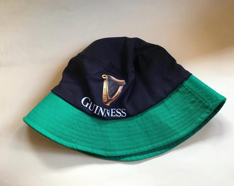 Op de afbeelding: Een zwarte en groene bucket hat met het Guinness-logo en een harpdesign. Het logo luidt "GUINNESS" in witte tekst.