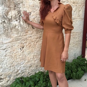May include: A woman wearing a brown dress with a ruffled neckline and puff sleeves. She is standing in front of a stone wall and has her hand on the wall. She is wearing sunglasses and has long red hair.