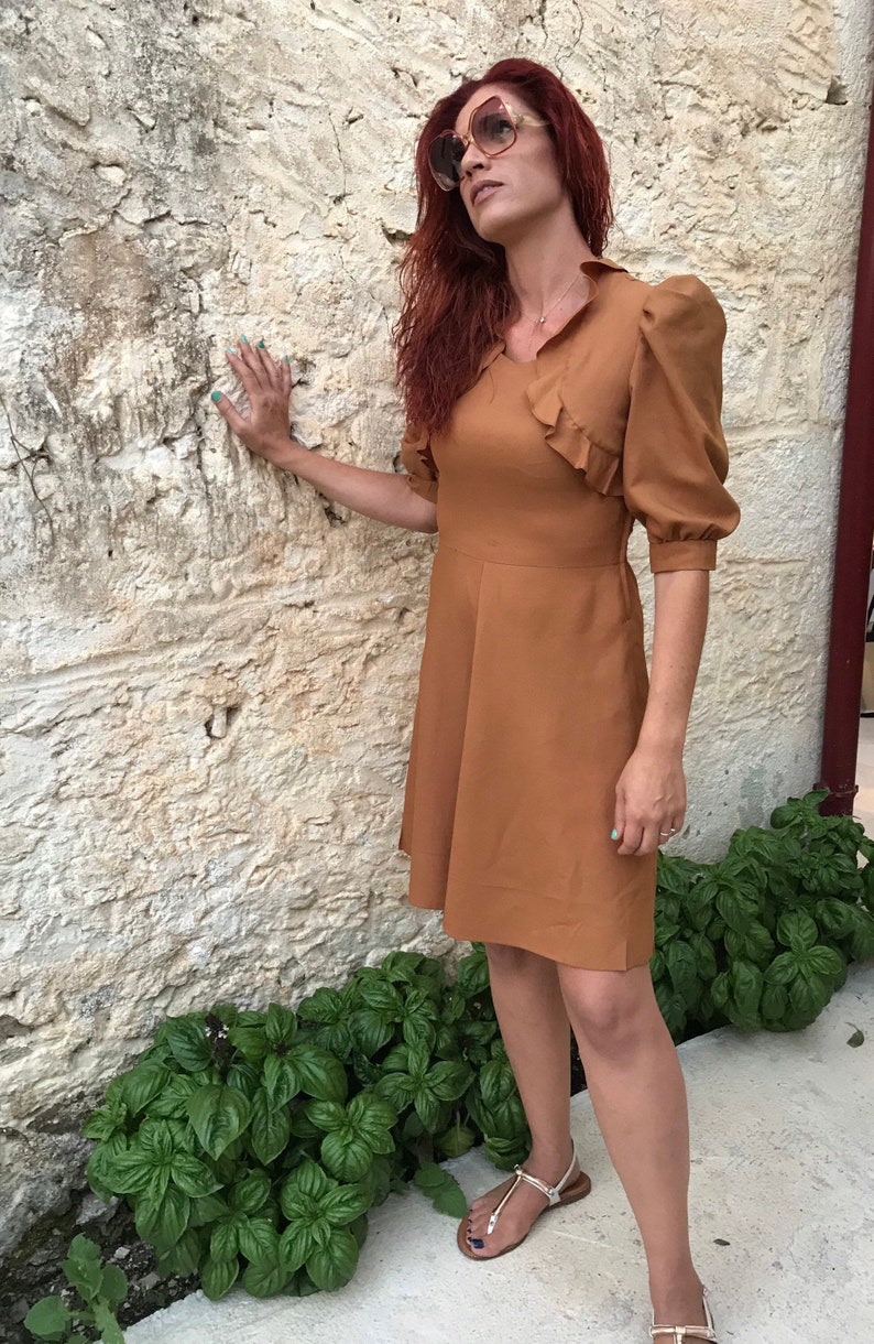 May include: A woman wearing a brown dress with a ruffled neckline and puff sleeves. She is standing in front of a stone wall and has her hand on the wall. She is wearing sunglasses and has long red hair.
