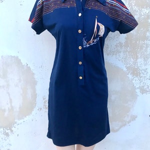 May include: A navy blue short-sleeved dress with a white sailboat graphic and gold buttons. The dress has a nautical-themed print on the sleeves with red, white, and blue stripes.