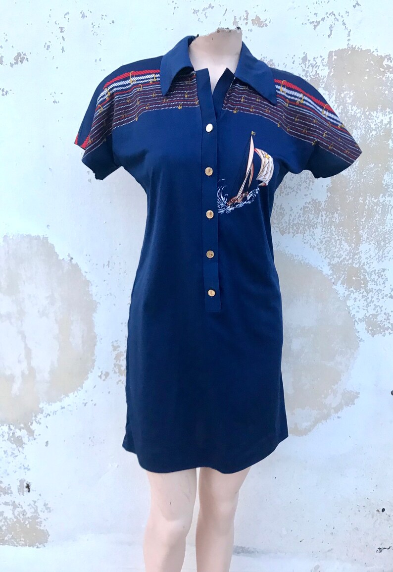 May include: A navy blue short-sleeved dress with a white sailboat graphic and gold buttons. The dress has a nautical-themed print on the sleeves with red, white, and blue stripes.