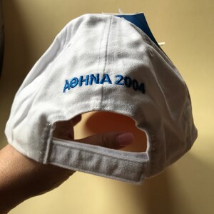 Puede incluir: Gorra de b&eacute;isbol blanca con texto bordado en azul que dice "ATENAS 2004".