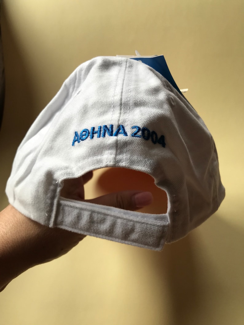 Puede incluir: Gorra de b&eacute;isbol blanca con texto bordado en azul que dice "ATENAS 2004".