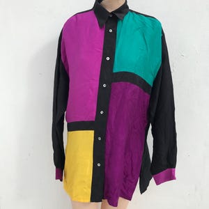 Könnte beinhalten: Ein langärmliges Button-Down-Hemd mit Farbblock-Design. Das Hemd hat Einsätze in Schwarz, Lila, Türkis und Gelb. Der Kragen und die Manschetten sind schwarz, und das Hemd ist zugeknöpft.