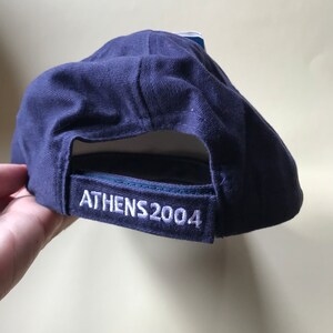 Puede incluir: Una gorra de b&eacute;isbol azul marino con costuras blancas y el texto "ATHENS 2004" bordado en la parte posterior.
