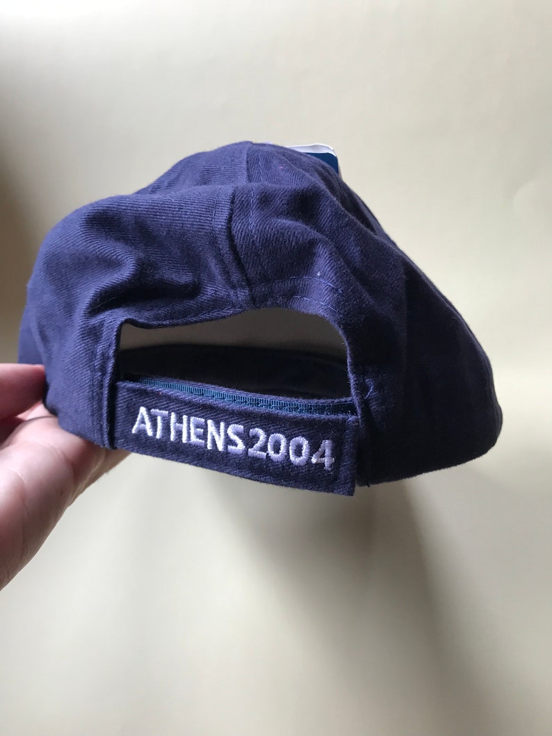 Puede incluir: Una gorra de b&eacute;isbol azul marino con costuras blancas y el texto "ATHENS 2004" bordado en la parte posterior.