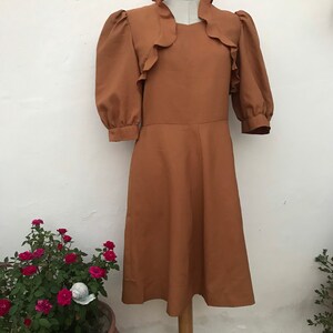 May include: A brown midi dress with a ruffled neckline and puffed sleeves. The dress has a fitted bodice and a flowing skirt.