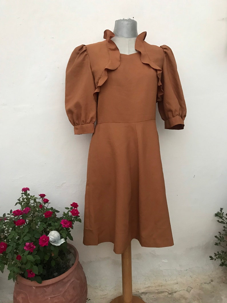May include: A brown midi dress with a ruffled neckline and puffed sleeves. The dress has a fitted bodice and a flowing skirt.