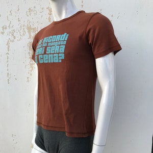 May include: Brown short-sleeved t-shirt with a blue graphic print that reads "Ti ricordi cosa hai mangiato ieri sera a cena?"