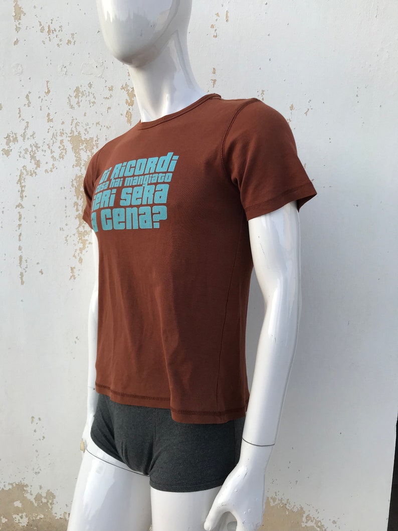 May include: Brown short-sleeved t-shirt with a blue graphic print that reads "Ti ricordi cosa hai mangiato ieri sera a cena?"