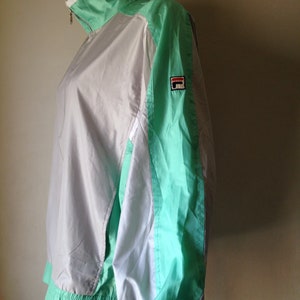 Puede incluir: Chaqueta deportiva vintage con dise&ntilde;o de bloques de color. La chaqueta presenta un cuerpo blanco y gris claro con detalles en verde menta en las mangas, el cuello y el dobladillo. Un peque&ntilde;o parche con el logotipo es visible en la manga superior izquierda. La chaqueta tiene cierre de cremallera.