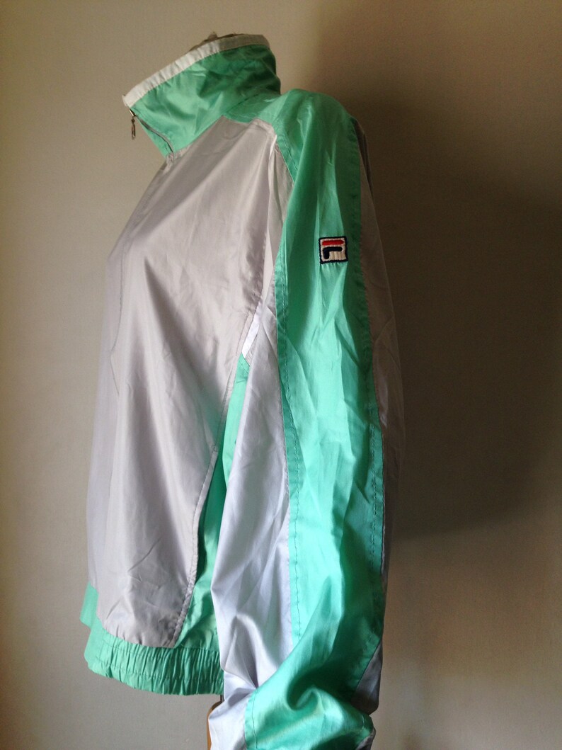 Puede incluir: Chaqueta deportiva vintage con dise&ntilde;o de bloques de color. La chaqueta presenta un cuerpo blanco y gris claro con detalles en verde menta en las mangas, el cuello y el dobladillo. Un peque&ntilde;o parche con el logotipo es visible en la manga superior izquierda. La chaqueta tiene cierre de cremallera.