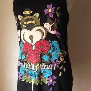 Op de afbeelding: Zwart tanktop met een kleurrijke grafische print van een hart met een sleutelgat, bloemen en een vogel. De tekst "key to my heart" is onder de print gedrukt.