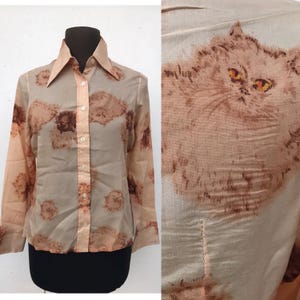 Könnte beinhalten: Ein Button-Down-Hemd mit Katzenmuster. Das Hemd hat einen hellpfirsichfarbenen Kragen und Manschetten mit einem Muster aus braunen und pfirsichfarbenen Katzenköpfen. Das Hemd wird auf einer schwarzen Schaufensterpuppe präsentiert.