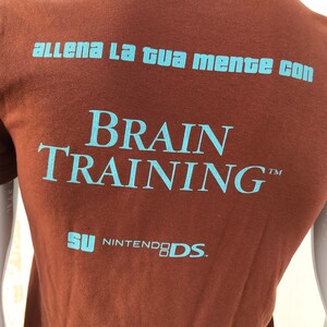 May include: Brown t-shirt with blue text that reads "allena la tua mente con BRAIN TRAINING&trade;  SU NINTENDO DS."