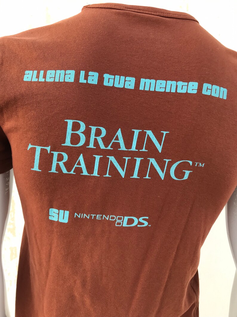 May include: Brown t-shirt with blue text that reads "allena la tua mente con BRAIN TRAINING&trade;  SU NINTENDO DS."
