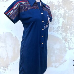 May include: A navy blue short-sleeved dress with a white, red and blue striped pattern on the shoulders and a sailboat graphic on the front. The dress has a collared neckline and button closure.