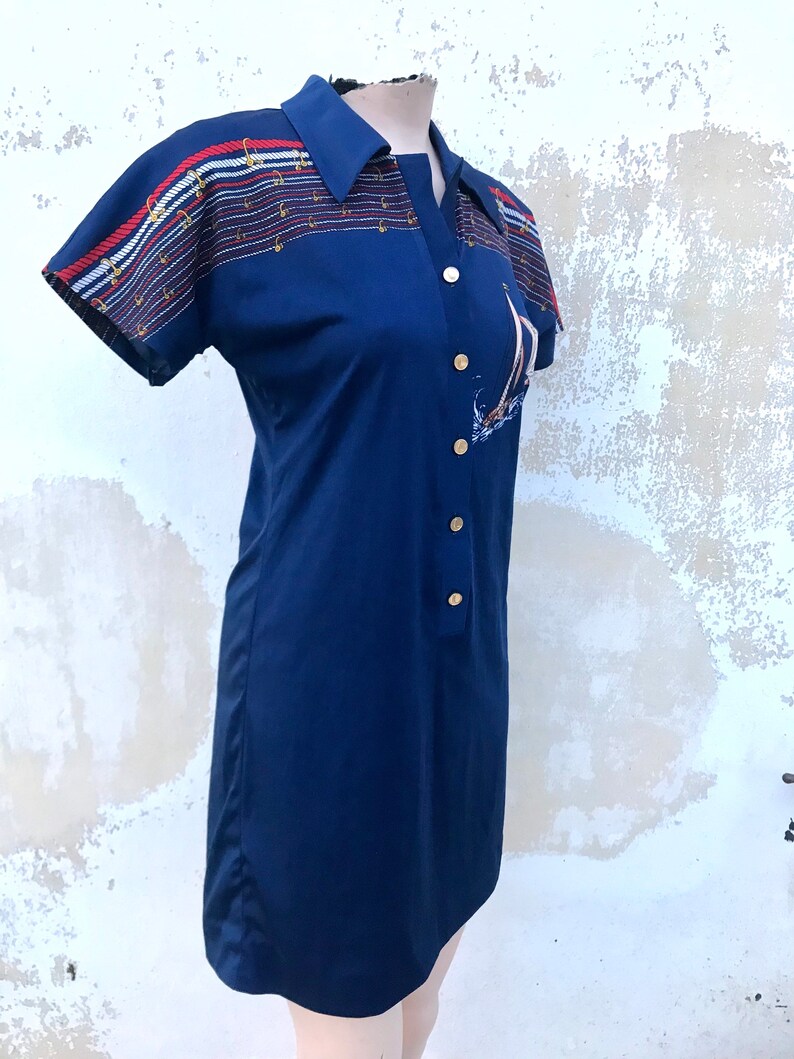 May include: A navy blue short-sleeved dress with a white, red and blue striped pattern on the shoulders and a sailboat graphic on the front. The dress has a collared neckline and button closure.