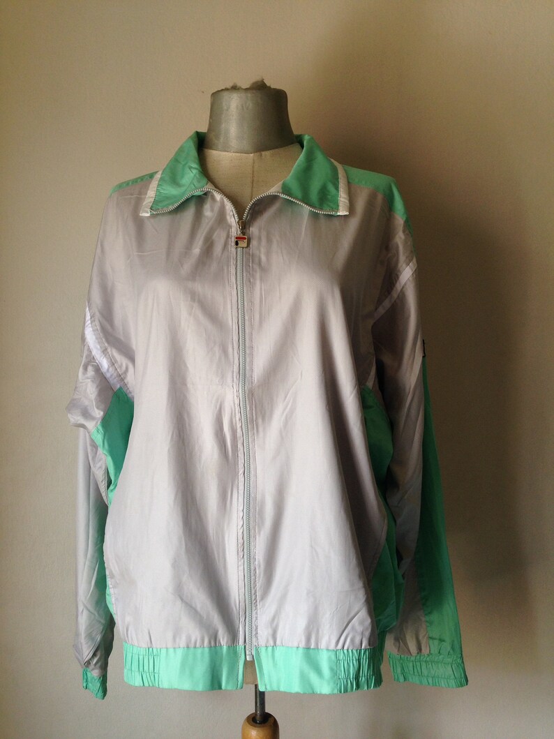 Puede incluir: Chaqueta deportiva vintage gris y verde menta con cierre de cremallera completo. La chaqueta presenta un cuello y detalles de manga blancos, y un dobladillo y pu&ntilde;os verde menta. La chaqueta se muestra en un maniqu&iacute;.