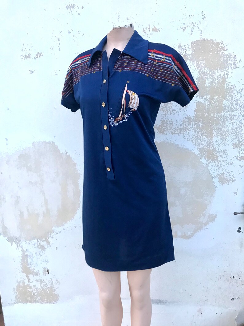 May include: A navy blue short-sleeved dress with a white sailboat graphic and a striped collar. The dress has gold buttons down the front.