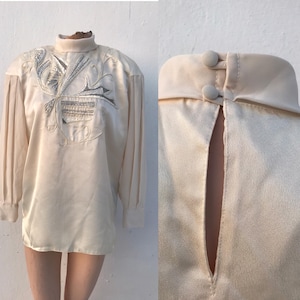 May include: Cream-colored blouse with a high, buttoned collar and long sleeves. The front features an embroidered design with silver and cream-colored thread. The sleeves have a pleated detail at the shoulder. The blouse is made of a silky material.