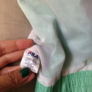Puede incluir: Primer plano del interior de una chaqueta Fila verde menta. La chaqueta presenta un forro de malla blanca y una etiqueta que dice "Made in Italy". El exterior de la chaqueta es de color verde menta liso.