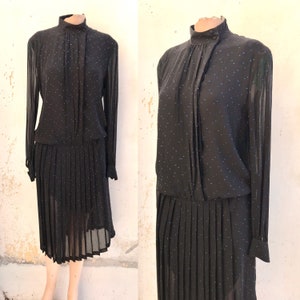 Può includere: Un abito nero in chiffon con collo alto e maniche lunghe. L'abito ha una gonna plissettata e un piccolo motivo a pois blu.