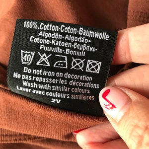 May include: Black care label with white text and symbols. The text reads "100% Cotton-Coton-Baumwolle Algod&oacute;n-Algod&atilde;o- Cotone-Katoen-&beta;&alpha;&mu;&beta;ά&kappa;&iota;- Puuvilla-Bomull Do not iron on decoration Ne pas repasser les d&eacute;corations Wash with similar colours Laver avec couleurs similaires 2V"
