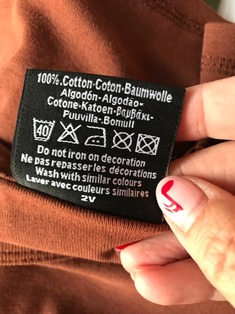 May include: Black care label with white text and symbols. The text reads "100% Cotton-Coton-Baumwolle Algod&oacute;n-Algod&atilde;o- Cotone-Katoen-&beta;&alpha;&mu;&beta;ά&kappa;&iota;- Puuvilla-Bomull Do not iron on decoration Ne pas repasser les d&eacute;corations Wash with similar colours Laver avec couleurs similaires 2V"