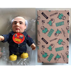 May include: A doll of a man with a bald head and gray hair, wearing a denim outfit and yellow shoes. The doll has a tag that says "GOABY AVARTI". Beside the doll is a box with a repeating pattern of the doll's image and the word "GOABY".
