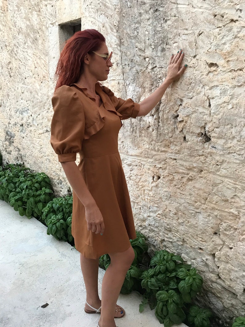 May include: A woman wearing a brown dress with a ruffled neckline and puff sleeves stands in front of a stone wall. She is touching the wall with her right hand.