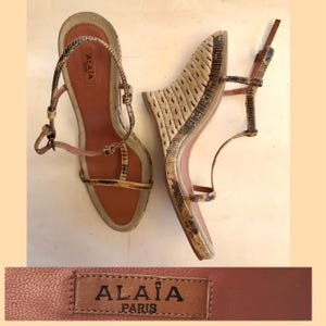 May include: A pair of Alaïa Paris wedge sandals featuring a snakeskin-patterned strap design with a T-strap silhouette, an adjustable ankle strap, and a textured wedge heel. The footbed is tan with the brand name Alaïa Paris printed on the heel.