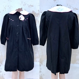 May include: Black dress with a white collar and pink trim. The dress has a button-up front and long sleeves. The dress is embroidered with small birds and the text "SIGOI" on the front.