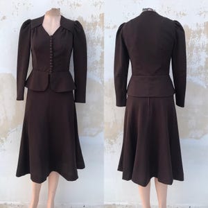 May include: A brown two-piece suit. The jacket has long sleeves, a V-neck, and a button closure. It is paired with a knee-length skirt. The suit has a vintage style with shoulder pads and a tailored fit. The skirt measures approximately 76 cm in length.