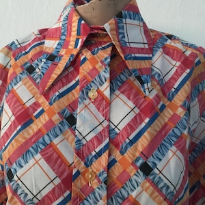 Puede incluir: Una camisa abotonada colorida con un vibrante patrón de cuadros. La camisa presenta una mezcla de rayas y cuadrados rojos, naranjas, azules y blancos. El cuello y los puños son visibles, y la camisa está abotonada. La tela parece ligera.