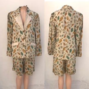 May include: A two-piece suit featuring a blazer and shorts. The blazer has a beige collar and pockets, with a button closure. The suit is patterned with green, brown, and yellow leaf and floral designs on a beige background. The shorts match the blazer's pattern.