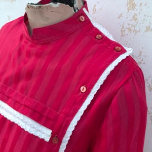 Könnte beinhalten: Eine leuchtend rote Bluse mit vertikalen Streifen und einem hohen Kragen. Die Bluse hat eine weiße Spitzenborte am Kragen und der Knopfleiste, mit passenden roten Knöpfen. Der Stoff scheint leicht und halbtransparent zu sein.