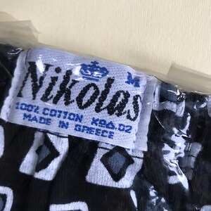 May include: A white tag with blue text and a crown logo reads "Nikolas 100% Cotton KBA. 02 Made in Greece". The tag is attached to black fabric with a white and grey geometric pattern.