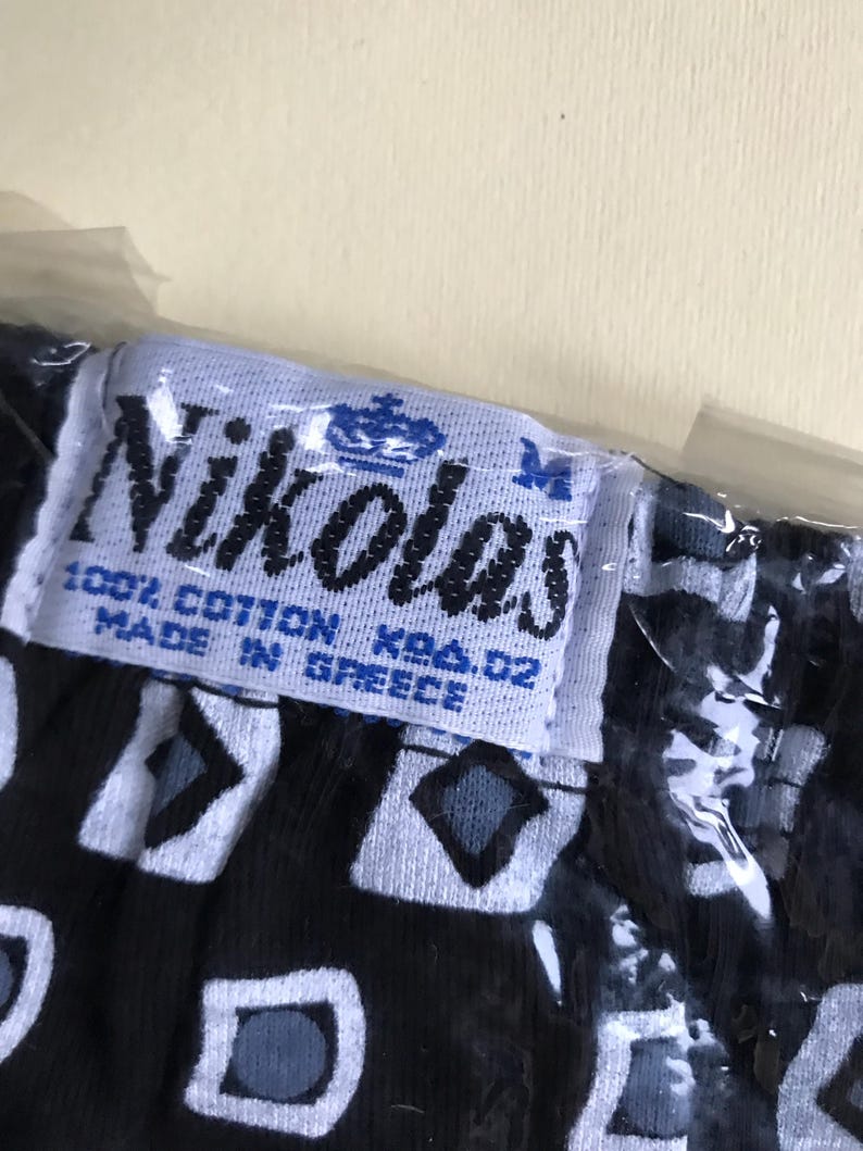 May include: A white tag with blue text and a crown logo reads "Nikolas 100% Cotton KBA. 02 Made in Greece". The tag is attached to black fabric with a white and grey geometric pattern.