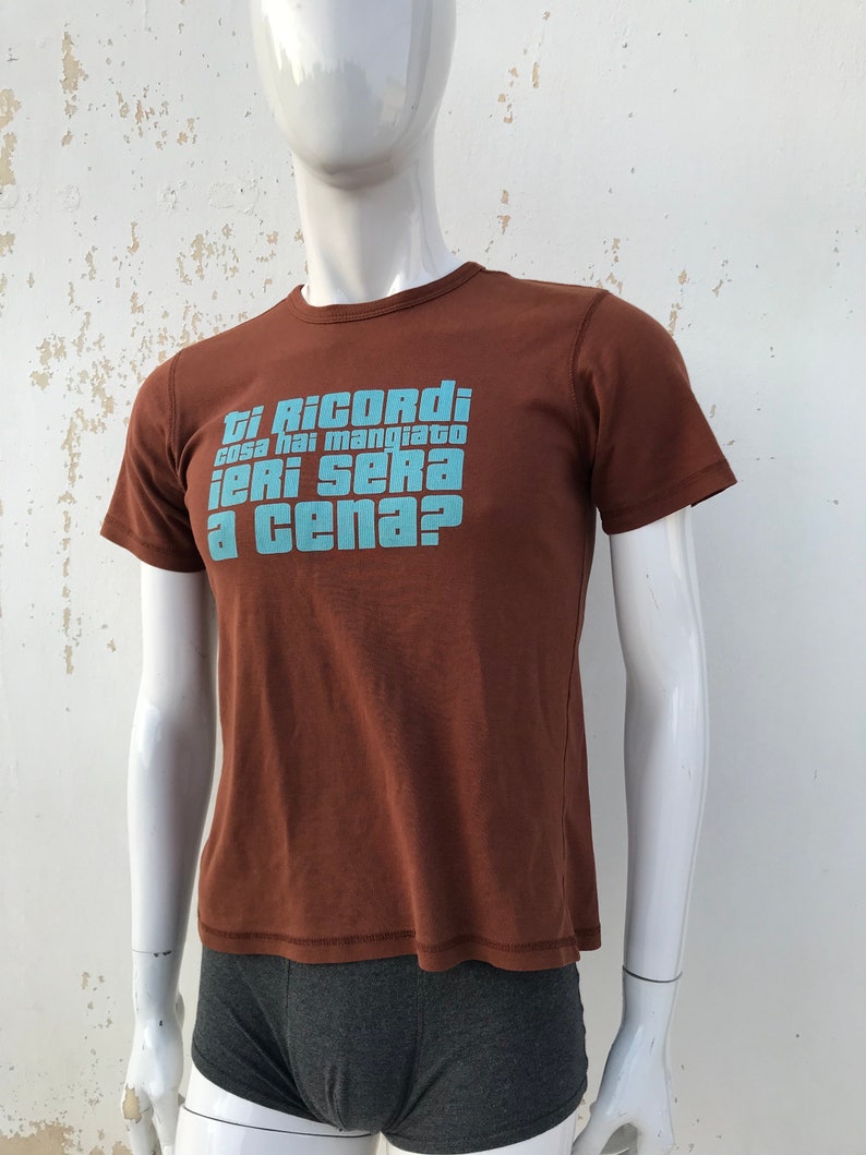May include: A brown t-shirt with a blue graphic that says "Ti Ricordi cosa hai mangiato ieri sera a cena?"