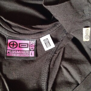 Op de afbeelding: Zwart T-shirt met een roze en zwart label met de tekst "Plusminus L Created by Chiemsee" en een wit label met een plus- en minteken.