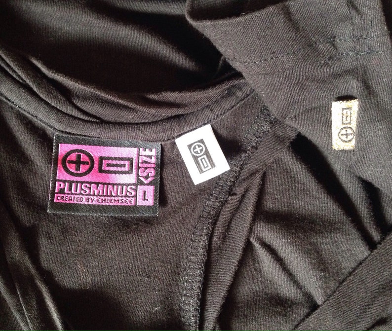 Op de afbeelding: Zwart T-shirt met een roze en zwart label met de tekst "Plusminus L Created by Chiemsee" en een wit label met een plus- en minteken.