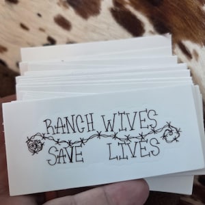 May include: A stack of white rectangular stickers with the text "RANCH WIVES SAVE LIVES" in black, with barbed wire and rose illustrations. The stickers are held in a hand.