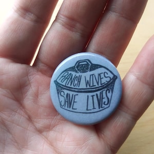 May include: A round, blue button with a black and white illustration of a pot with the text "Ranch Wives Save Lives".