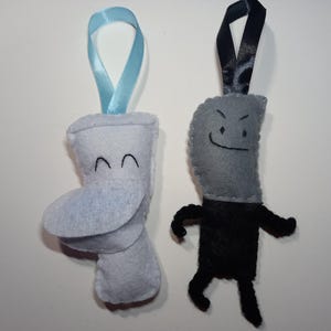 May include: Two handmade felt ornaments. One is a light blue toilet with a smiling face and a blue ribbon. The other is a grey knife with a smiling face and black arms and legs, with a black ribbon.