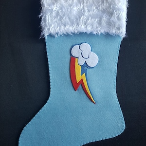 May include: A blue felt Christmas stocking with a white faux fur cuff. The stocking features a felt applique of Rainbow Dash's cutie mark, a rainbow with a cloud and lightning bolt.